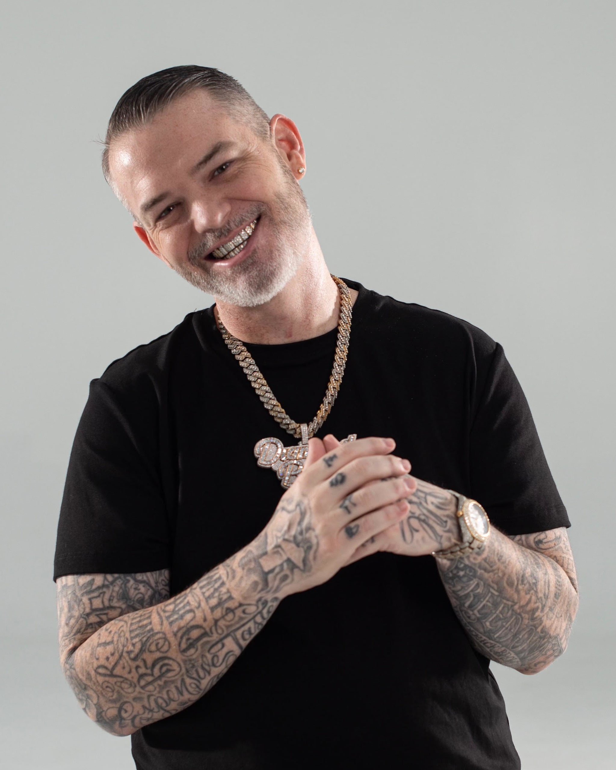 A man with tattoos and a beard smiles, wearing a black shirt, gold chains, and a watch.